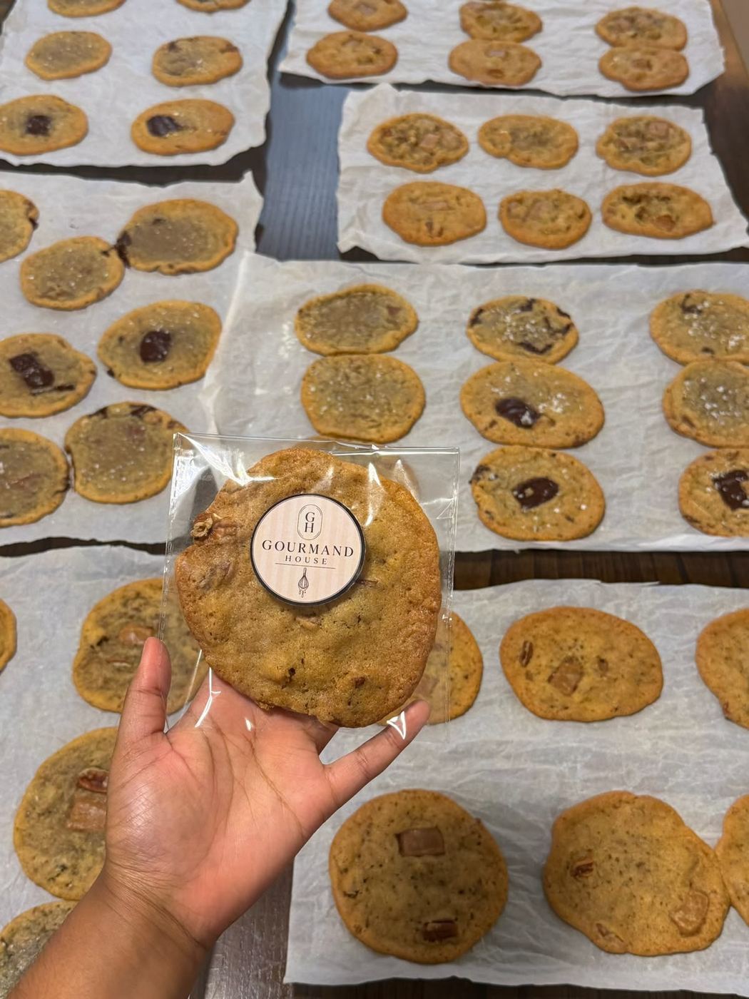 A hand holding a Gourmand House cookie sealed with the round logo sticker, over trays of freshly baked cookies